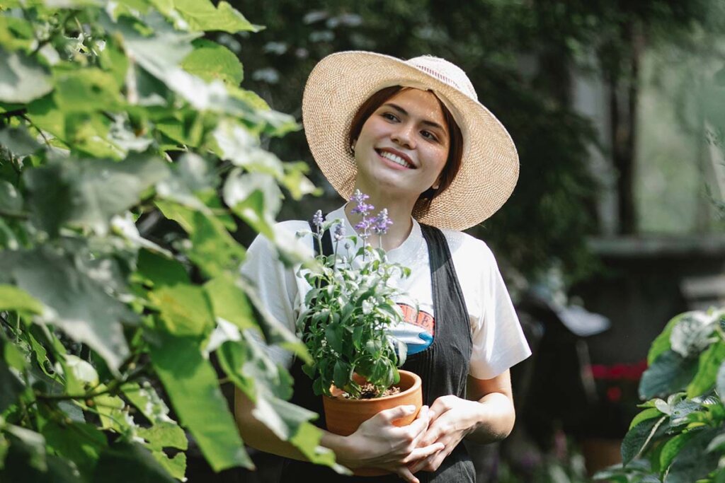 Eine Frau in gruner Latzhose, weißem Shirt, mit Hut haelt einen Topf Lavendel in der Hand und geht durch einen Garten.