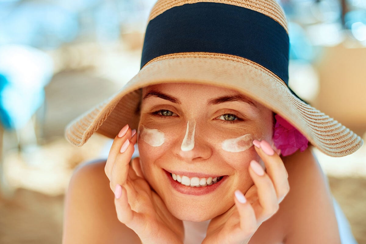 Frau mit Sonnenhut und Sonnencreme im Gesicht laechelt in die Kamera.